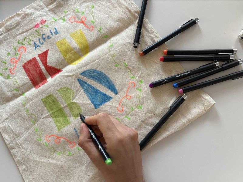auf dem Bild siehst du eine Hand, welche einen Stift in der Hand hält und einen Jutebeutel mit dem Logo vom KUBA gestaltet