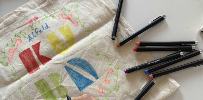 auf dem Bild siehst du eine Hand, welche einen Stift in der Hand hält und einen Jutebeutel mit dem Logo vom KUBA gestaltet